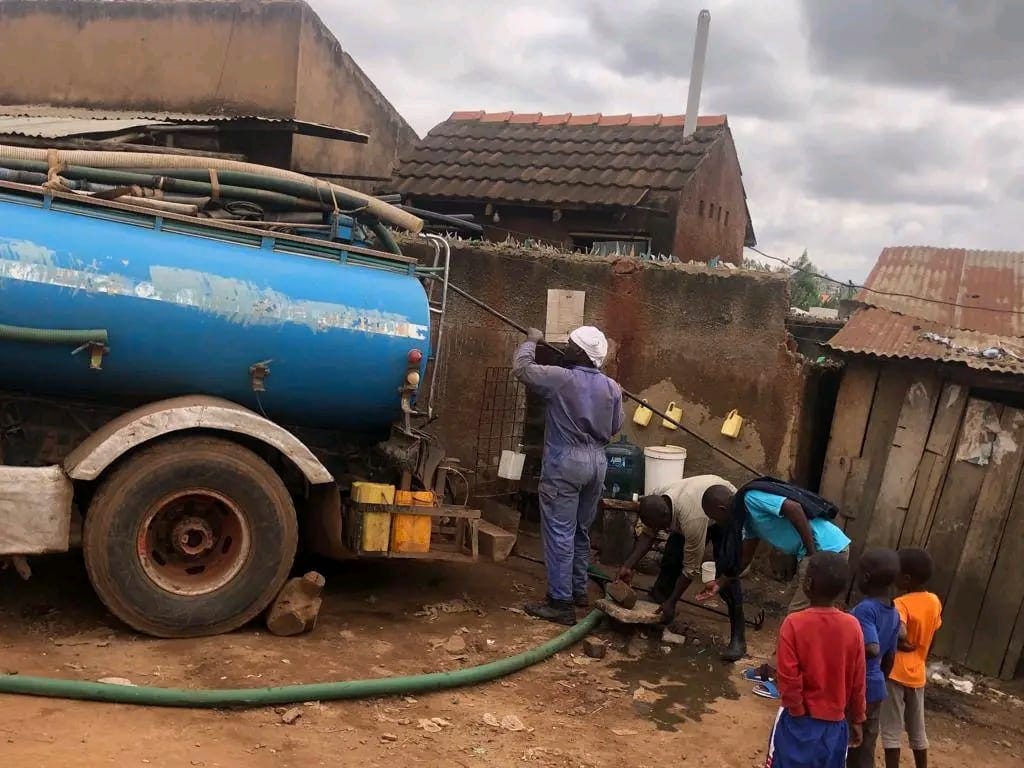 Workers emptying a shared community latrine with a hose and sanitation truck as residents watch